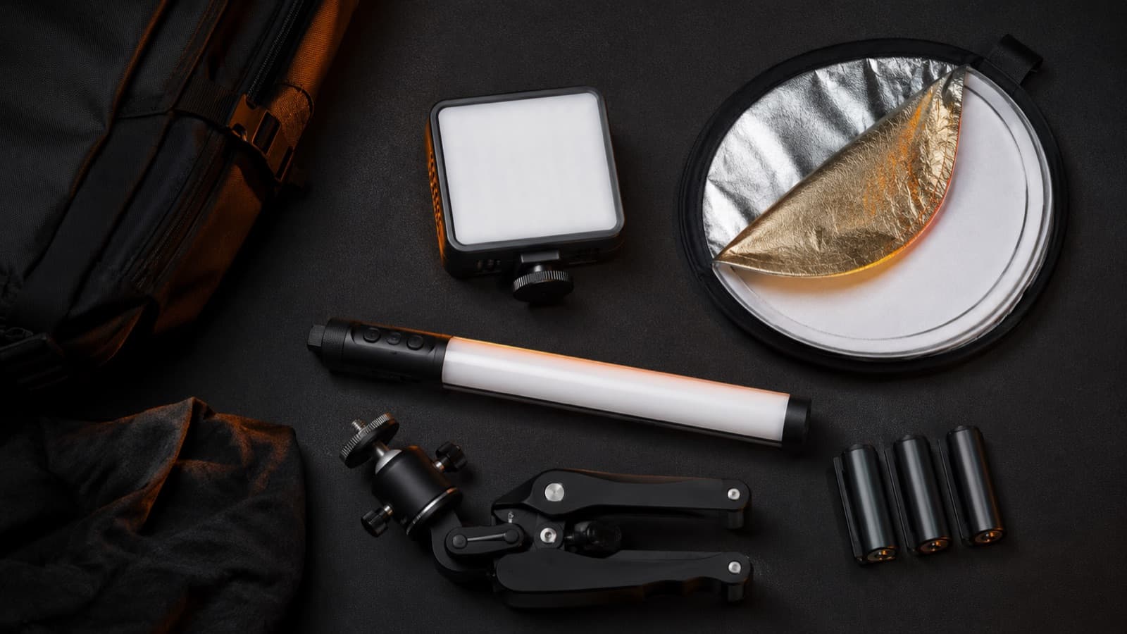 Flat-lay de um kit de luz leve: painel pequeno, rebatedor dobrado, tubo RGB, mini tripé e baterias